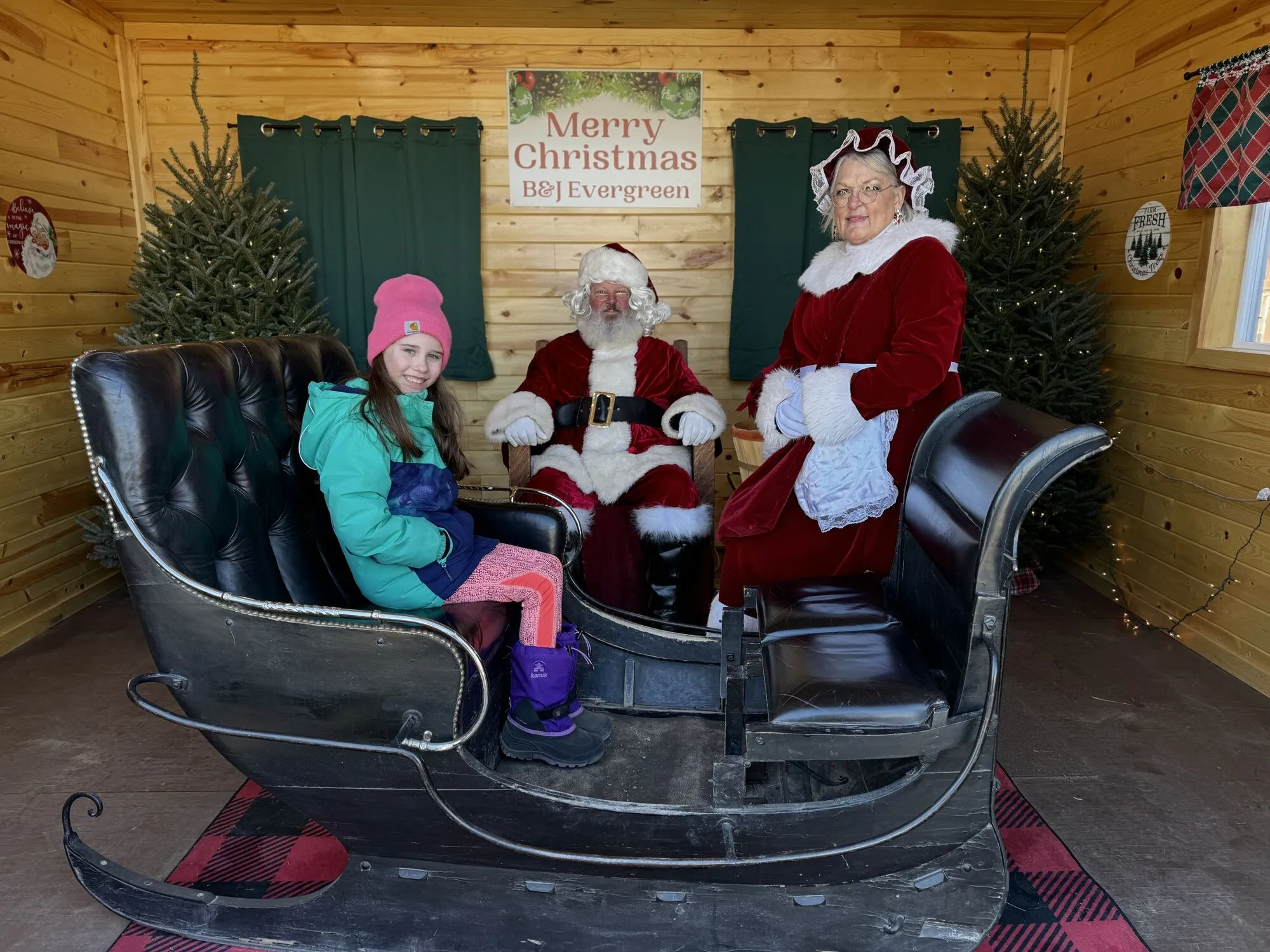 A girl sitting in the full-sized sleigh with Santa and Mrs. Claus in the cabin at the Clear Lake farm